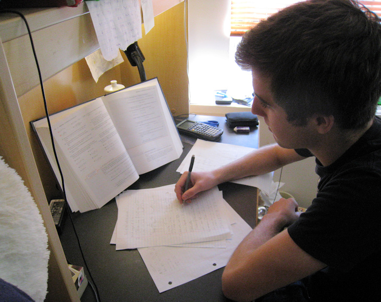Young man sitting at a desk with a book propped open on it.  He is writing notes on sheets of paper.